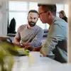 Zwei Männer arbeiten in einem modernen Büro an einem Schreibtisch mit Laptops zusammen; im Hintergrund ist ein verschwommener Kollege zu sehen.
