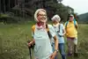 Drei Senioren wandern auf einem grasbewachsenen Weg, lächeln und tragen Rucksäcke. Im Hintergrund sind Bäume und Hügel zu sehen.