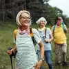 Drei Senioren wandern auf einem grasbewachsenen Weg, lächeln und tragen Rucksäcke. Im Hintergrund sind Bäume und Hügel zu sehen.