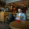 Man sits in a cozy cafe, holding a cup of coffee and looking at his phone.