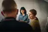 A woman in a green sweater smiles warmly while talking to a person in the foreground, with another person in the background, in a casual setting.